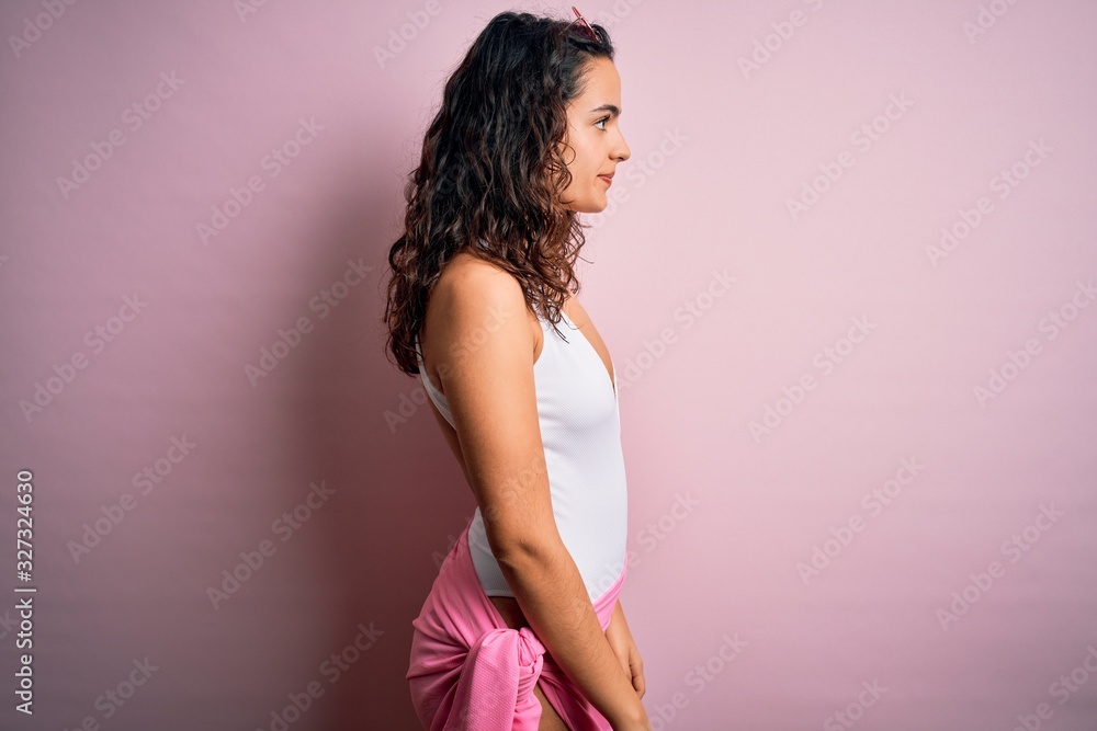 Beautiful woman with curly hair on vacation wearing white swimsuit over pink background looking to side, relax profile pose with natural face and confident smile. Beautiful woman with curly hair on vacation wearing white swimsuit over pink background looking to side, relax profile pose with natural face and confident smile.