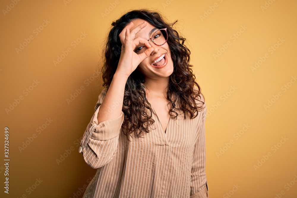 Beautiful woman with curly hair wearing striped shirt and glasses over yellow background doing ok gesture with hand smiling, eye looking through fingers with happy face. Beautiful woman with curly hair wearing striped shirt and glasses over yellow background doing ok gesture with hand smiling, eye looking through fingers with happy face.