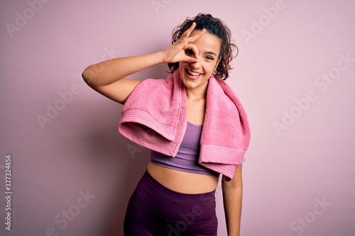 Young beautiful sportswoman with curly hair doing sport using towel over pink background doing ok gesture with hand smiling, eye looking through fingers with happy face.