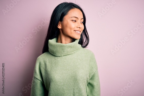 Young beautiful chinese woman wearing turtleneck sweater over isolated pink background looking away to side with smile on face, natural expression. Laughing confident.