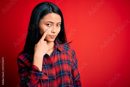 Young beautiful chinese woman wearing casual shirt over isolated red background Pointing to the eye watching you gesture, suspicious expression © Krakenimages.com