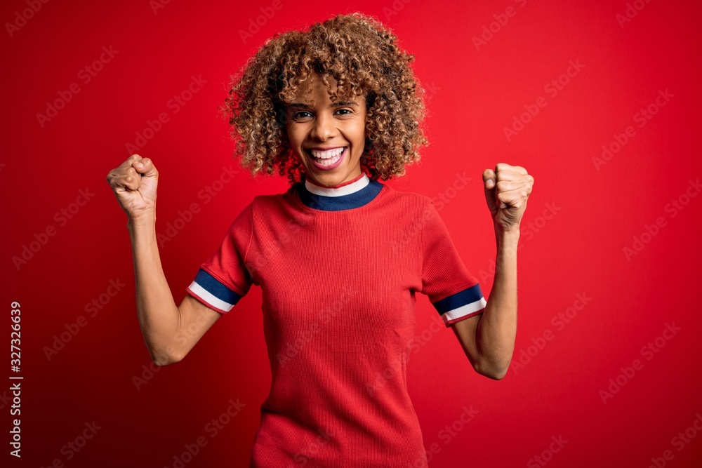 Young beautiful african american woman wearing casual t-shirt standing over red background celebrating surprised and amazed for success with arms raised and open eyes. Winner concept. Young beautiful african american woman wearing casual t-shirt standing over red background celebrating surprised and amazed for success with arms raised and open eyes. Winner concept.