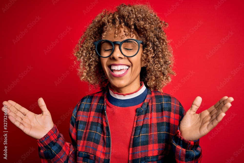 Young beautiful african american woman wearing casual shirt and glasses over red background celebrating mad and crazy for success with arms raised and closed eyes screaming excited. Winner concept