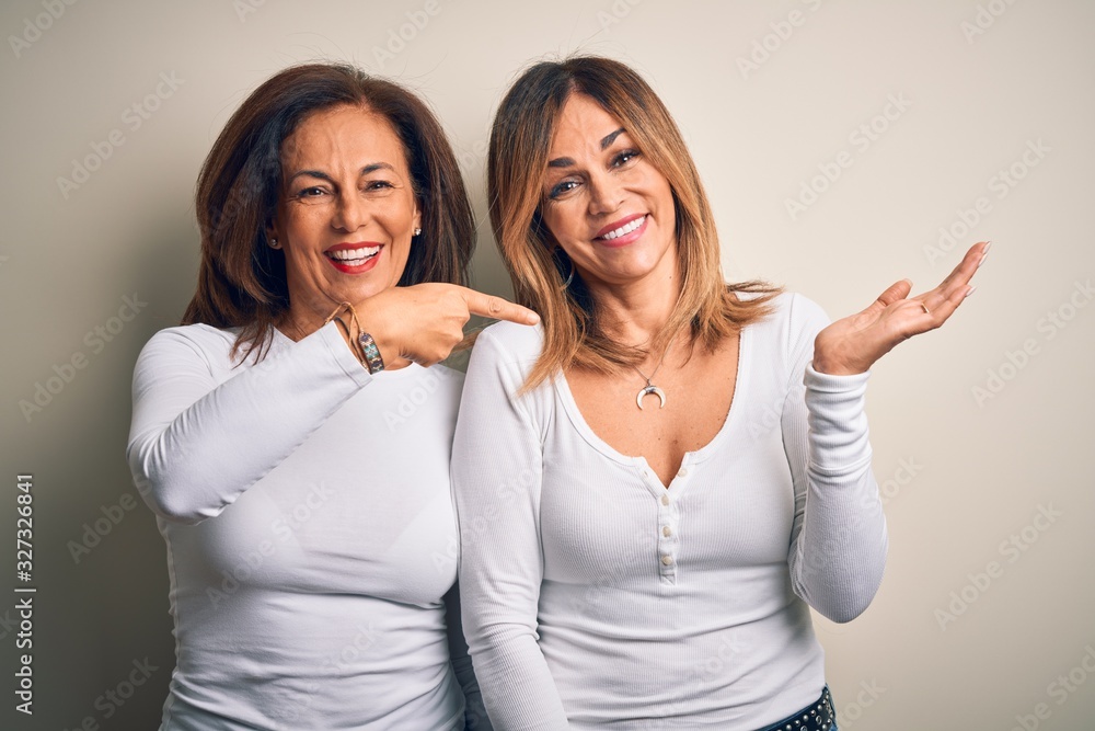 Middle age beautiful couple of sisters wearing casual t-shirt over isolated white background amazed and smiling to the camera while presenting with hand and pointing with finger. Middle age beautiful couple of sisters wearing casual t-shirt over isolated white background amazed and smiling to the camera while presenting with hand and pointing with finger.