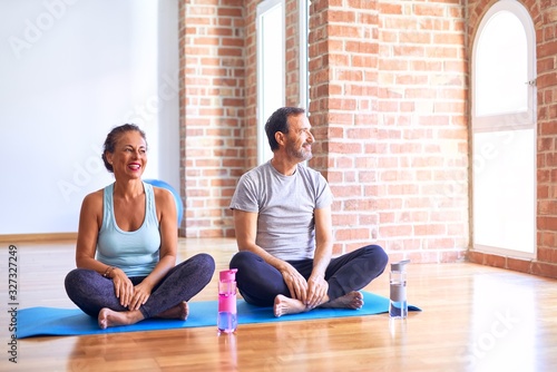 Middle age sporty couple sitting on mat doing stretching yoga exercise at gym looking away to side with smile on face, natural expression. Laughing confident.