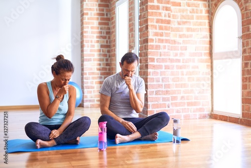 Middle age sporty couple sitting on mat doing stretching yoga exercise at gym feeling unwell and coughing as symptom for cold or bronchitis. Healthcare concept.