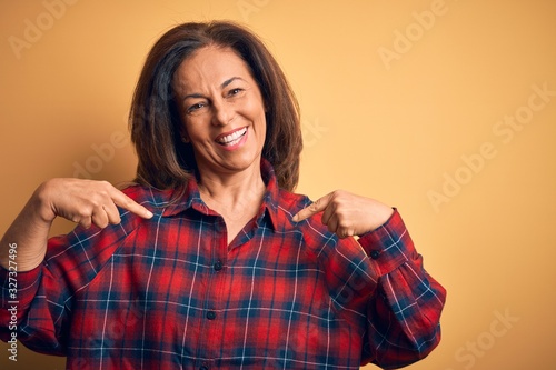 Middle age beautiful woman wearing casual shirt standing over isolated yellow background looking confident with smile on face, pointing oneself with fingers proud and happy.