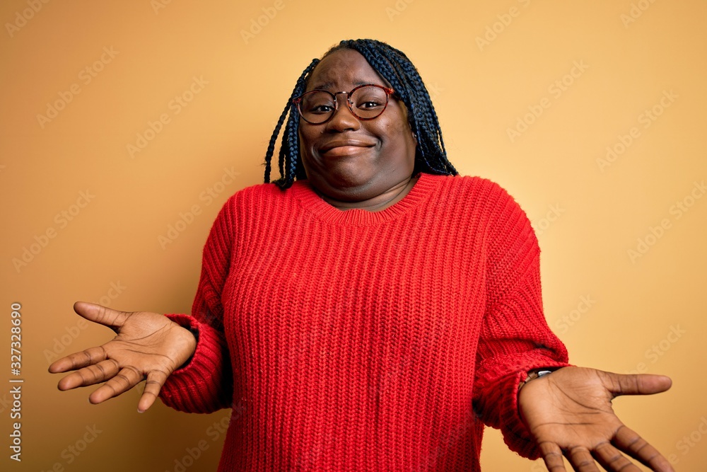 African american plus size woman with braids wearing casual sweater over yellow background clueless and confused expression with arms and hands raised. Doubt concept. African american plus size woman with braids wearing casual sweater over yellow background clueless and confused expression with arms and hands raised. Doubt concept.