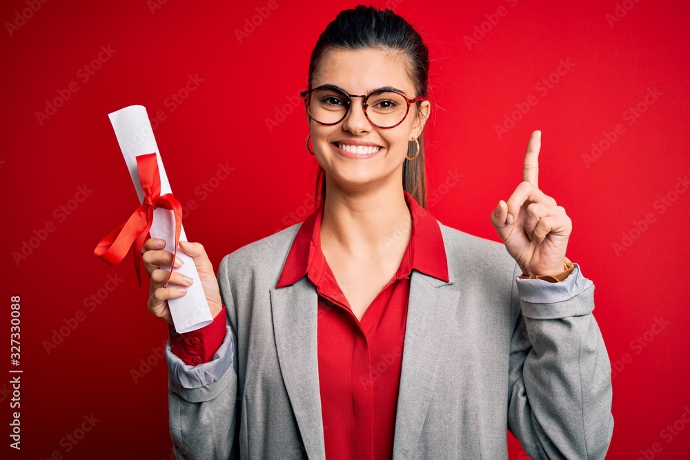 Young beautiful brunette student woman wearing glasses holding university degree diploma surprised with an idea or question pointing finger with happy face, number one