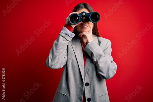 Young beautiful woman with blue eyes using binoculars over isolated red background serious face thinking about question, very confused idea