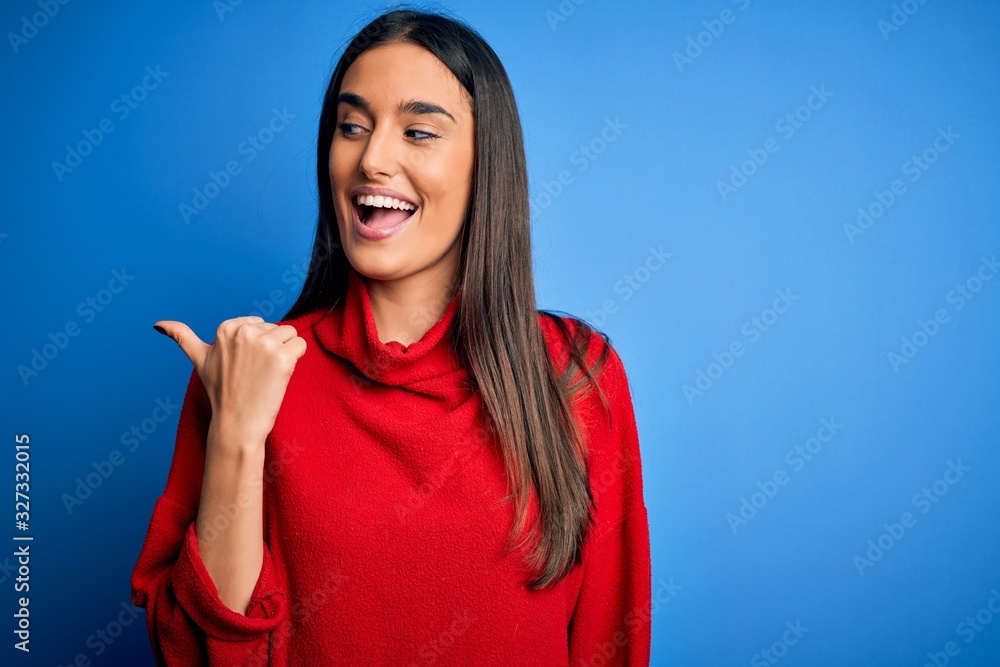 Young beautiful brunette woman wearing red casual sweater over isolated blue background smiling with happy face looking and pointing to the side with thumb up. Young beautiful brunette woman wearing red casual sweater over isolated blue background smiling with happy face looking and pointing to the side with thumb up.