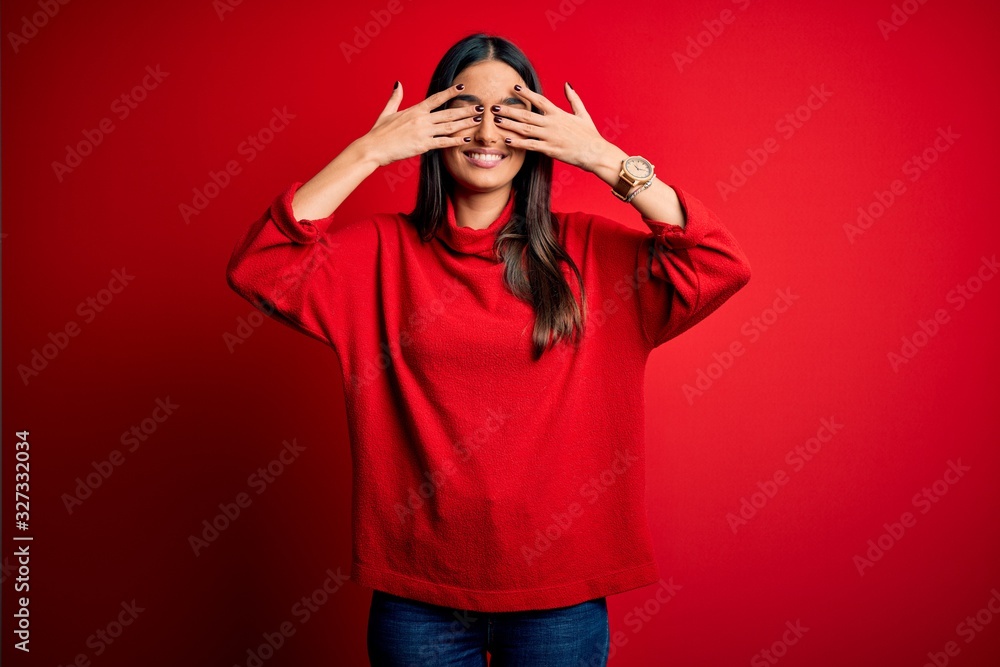 Young beautiful brunette woman wearing casual sweater over isolated red background covering eyes with hands smiling cheerful and funny. Blind concept. Young beautiful brunette woman wearing casual sweater over isolated red background covering eyes with hands smiling cheerful and funny. Blind concept.