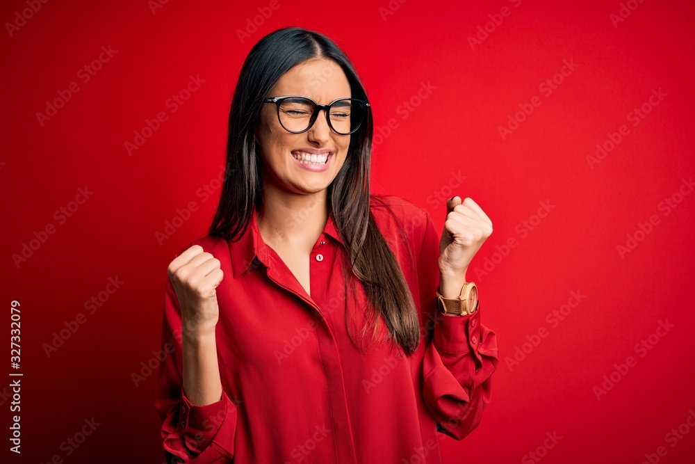 Young beautiful brunette woman wearing casual shirt and glasses over red background very happy and excited doing winner gesture with arms raised, smiling and screaming for success. Celebration Young beautiful brunette woman wearing casual shirt and glasses over red background very happy and excited doing winner gesture with arms raised, smiling and screaming for success. Celebration