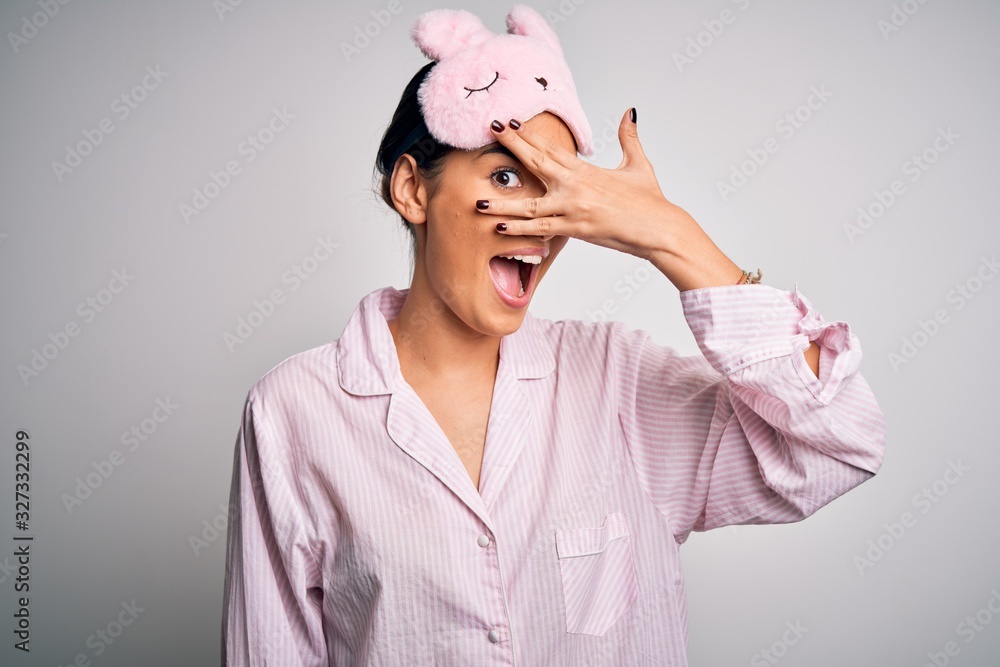 Young beautiful brunette woman wearing pajama and sleep mask over white background peeking in shock covering face and eyes with hand, looking through fingers with embarrassed expression. Young beautiful brunette woman wearing pajama and sleep mask over white background peeking in shock covering face and eyes with hand, looking through fingers with embarrassed expression.