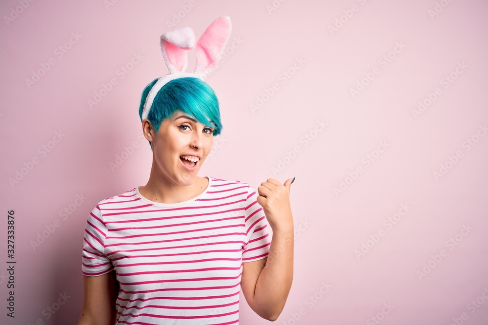 Young woman with fashion blue hair wearing easter rabbit ears over pink background smiling with happy face looking and pointing to the side with thumb up. Young woman with fashion blue hair wearing easter rabbit ears over pink background smiling with happy face looking and pointing to the side with thumb up.