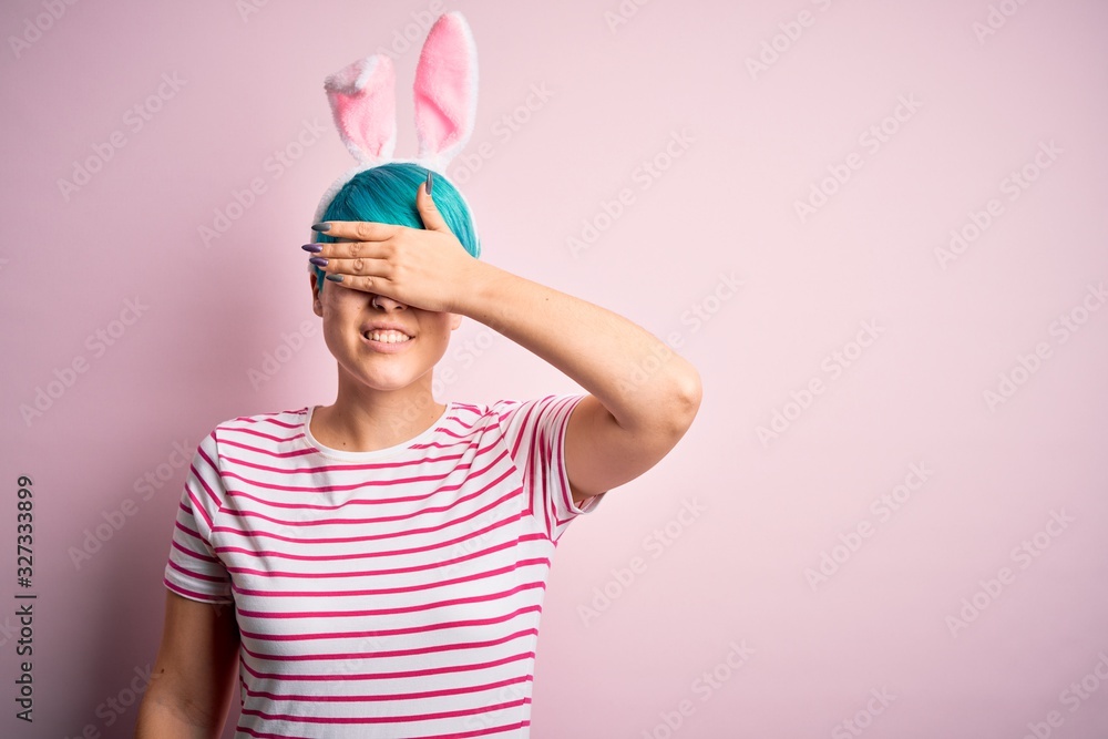 Young woman with fashion blue hair wearing easter rabbit ears over pink background smiling and laughing with hand on face covering eyes for surprise. Blind concept. Young woman with fashion blue hair wearing easter rabbit ears over pink background smiling and laughing with hand on face covering eyes for surprise. Blind concept.