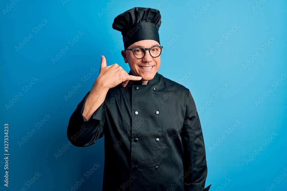 Middle age handsome grey-haired chef man wearing cooker uniform and hat smiling doing phone gesture with hand and fingers like talking on the telephone. Communicating concepts. Middle age handsome grey-haired chef man wearing cooker uniform and hat smiling doing phone gesture with hand and fingers like talking on the telephone. Communicating concepts.