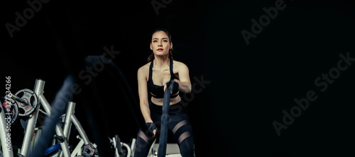 wide banner with copy space of athletic asian strong woman slim body training and working out with battle rope at fitness gym in dark background, bodybuilder, exercise fitness, workout, sport concept