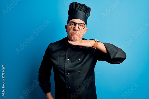 Middle age handsome grey-haired chef man wearing cooker uniform and hat cutting throat with hand as knife, threaten aggression with furious violence