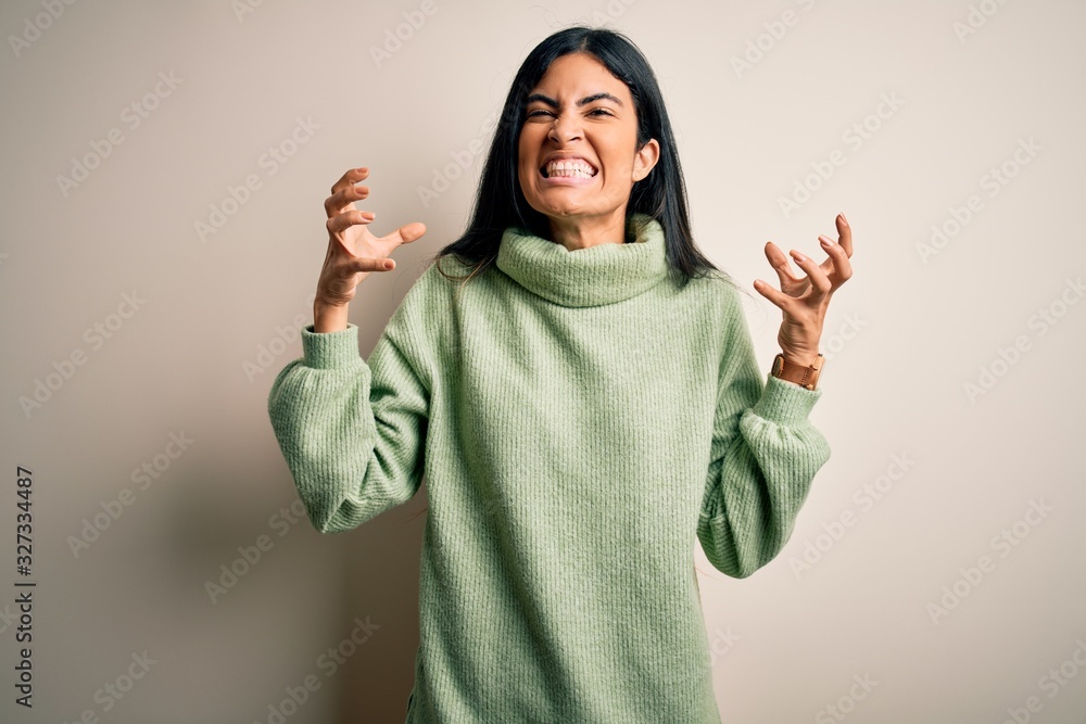 Young beautiful hispanic woman wearing green winter sweater over isolated background Shouting frustrated with rage, hands trying to strangle, yelling mad Young beautiful hispanic woman wearing green winter sweater over isolated background Shouting frustrated with rage, hands trying to strangle, yelling mad
