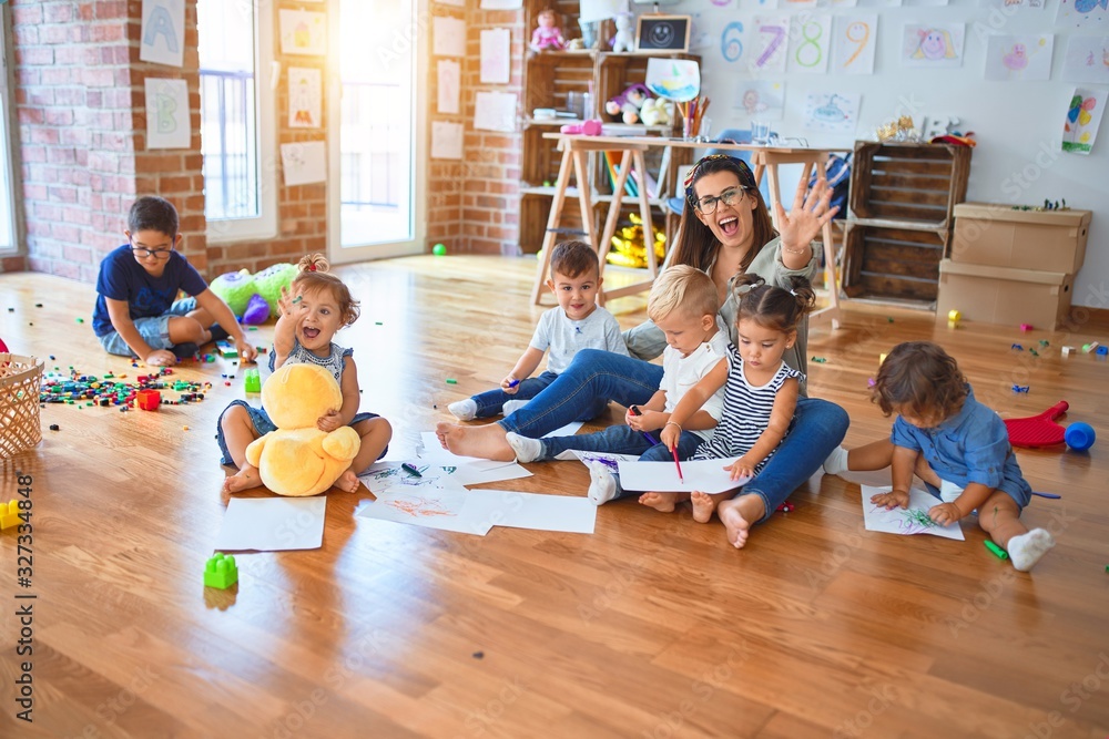 Beautiful teacher and group of toddlers sitting on the floor drawing using paper and pencil around lots of toys at kindergarten - obrazy, fototapety, plakaty 