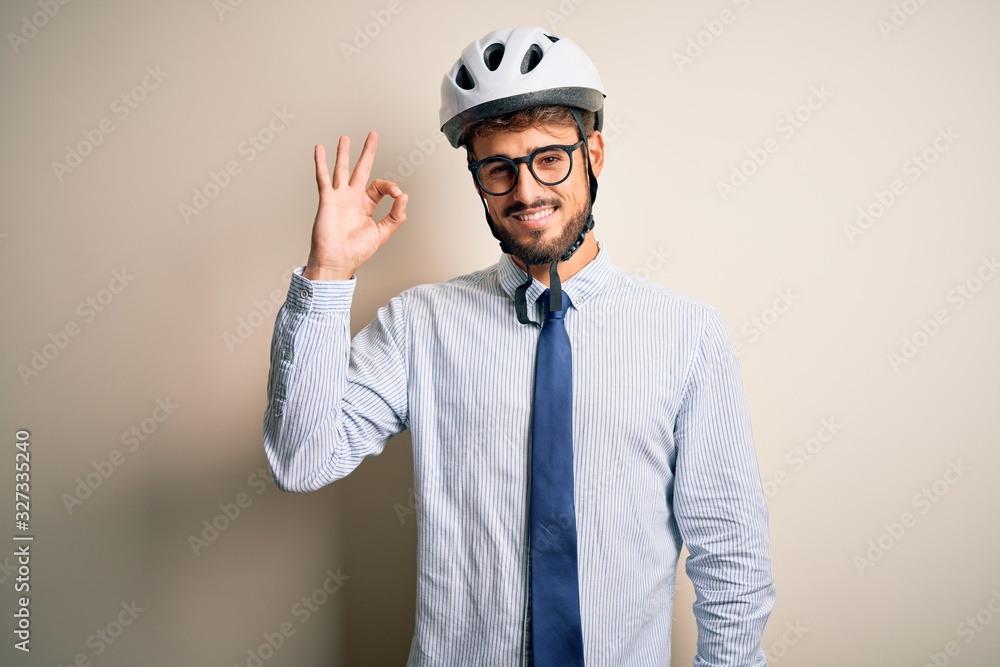 Young businessman wearing glasses and bike helmet standing over isolated white bakground smiling positive doing ok sign with hand and fingers. Successful expression. Young businessman wearing glasses and bike helmet standing over isolated white bakground smiling positive doing ok sign with hand and fingers. Successful expression.