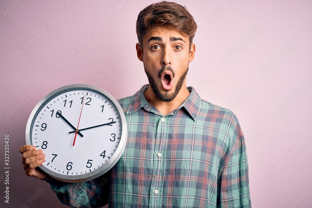 Young man with beard doing countdown using big clock over isolated pink background scared in shock with a surprise face, afraid and excited with fear expression Young man with beard doing countdown using big clock over isolated pink background scared in shock with a surprise face, afraid and excited with fear expression