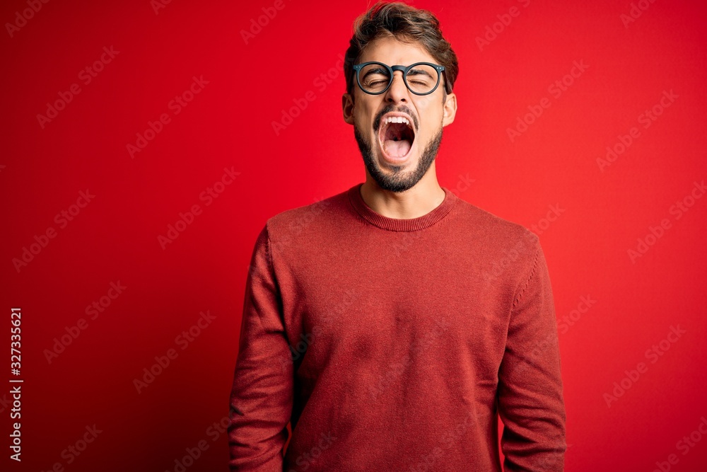Obraz premium Young handsome man with beard wearing glasses and sweater standing over red background angry and mad screaming frustrated and furious, shouting with anger. Rage and aggressive concept.