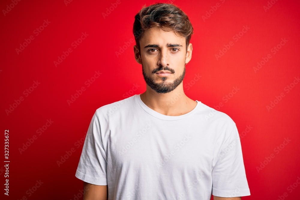 Young handsome man with beard wearing casual t-shirt standing over red background with serious expression on face. Simple and natural looking at the camera. Young handsome man with beard wearing casual t-shirt standing over red background with serious expression on face. Simple and natural looking at the camera.