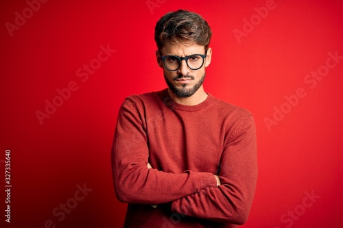 Young handsome man with beard wearing glasses and sweater standing over red background skeptic and nervous, disapproving expression on face with crossed arms. Negative person. © Krakenimages.com