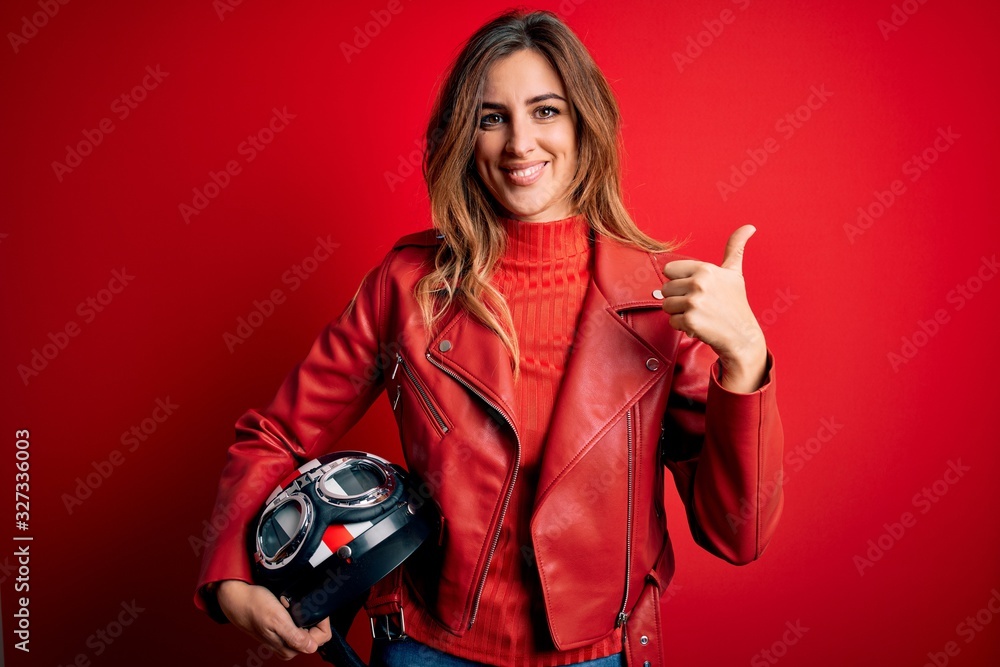 Beautiful brunette motorcyclist woman wearing red jacket holding motorcycle helmet happy with big smile doing ok sign, thumb up with fingers, excellent sign Beautiful brunette motorcyclist woman wearing red jacket holding motorcycle helmet happy with big smile doing ok sign, thumb up with fingers, excellent sign