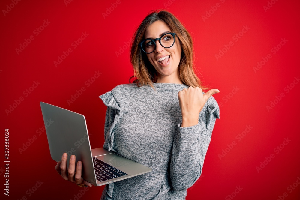 Naklejka premium Young beautiful brunette businesswoman wearing glasses using laptop over red background pointing and showing with thumb up to the side with happy face smiling