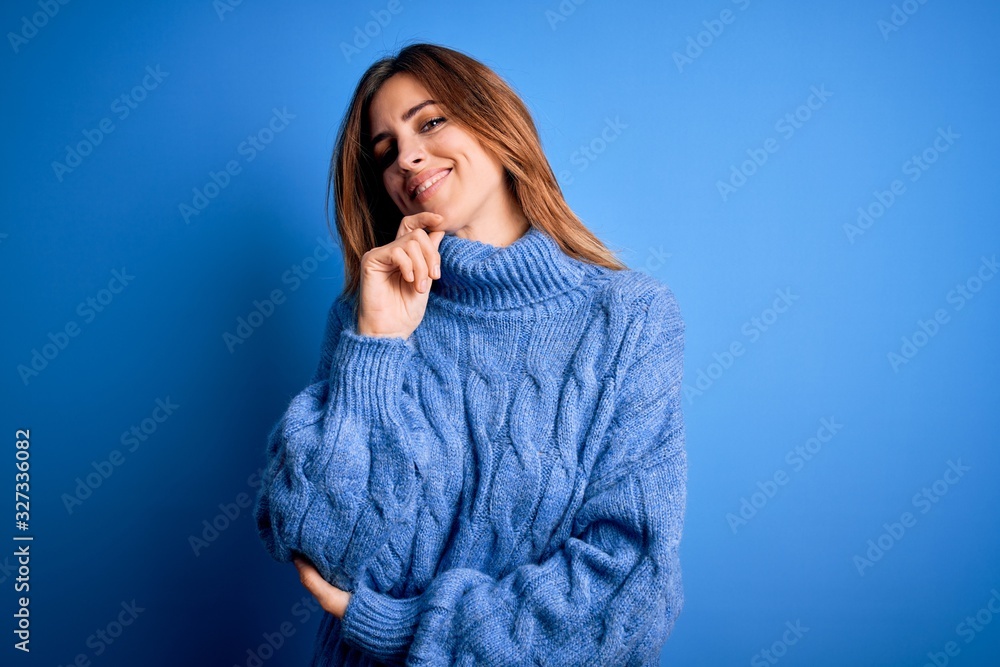 Young beautiful brunette woman wearing casual turtleneck sweater over blue background looking confident at the camera with smile with crossed arms and hand raised on chin. Thinking positive. Young beautiful brunette woman wearing casual turtleneck sweater over blue background looking confident at the camera with smile with crossed arms and hand raised on chin. Thinking positive.