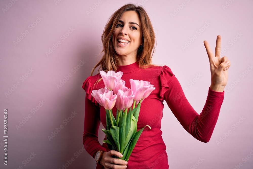 Young beautiful brunette woman holding bouquet of pink tulips over isolated background smiling looking to the camera showing fingers doing victory sign. Number two.