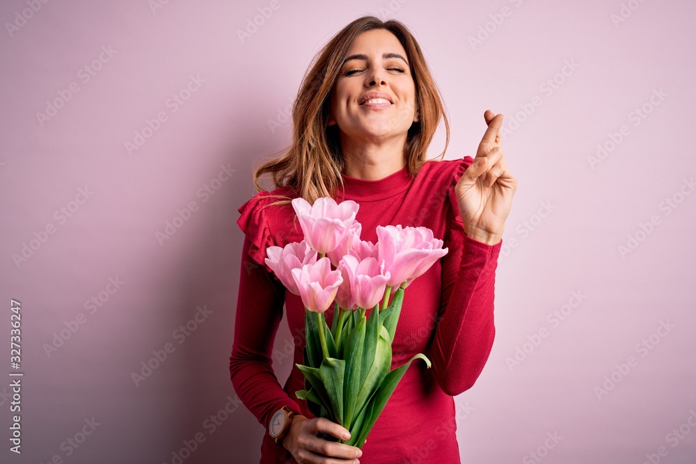 Young beautiful brunette woman holding bouquet of pink tulips over isolated background gesturing finger crossed smiling with hope and eyes closed. Luck and superstitious concept. Young beautiful brunette woman holding bouquet of pink tulips over isolated background gesturing finger crossed smiling with hope and eyes closed. Luck and superstitious concept.