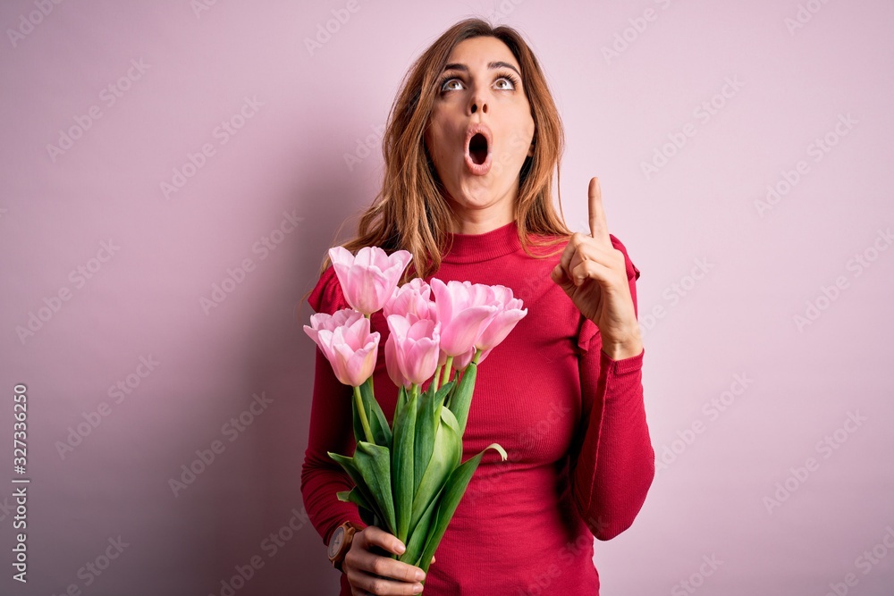 Young beautiful brunette woman holding bouquet of pink tulips over isolated background amazed and surprised looking up and pointing with fingers and raised arms. Young beautiful brunette woman holding bouquet of pink tulips over isolated background amazed and surprised looking up and pointing with fingers and raised arms.