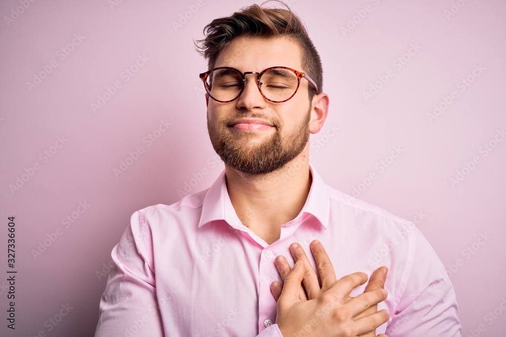 Young handsome blond man with beard and blue eyes wearing pink shirt and glasses smiling with hands on chest with closed eyes and grateful gesture on face. Health concept. Young handsome blond man with beard and blue eyes wearing pink shirt and glasses smiling with hands on chest with closed eyes and grateful gesture on face. Health concept.
