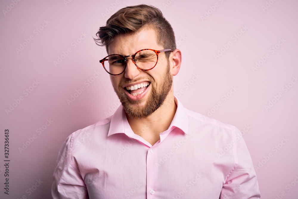 Young handsome blond man with beard and blue eyes wearing pink shirt and glasses winking looking at the camera with sexy expression, cheerful and happy face.