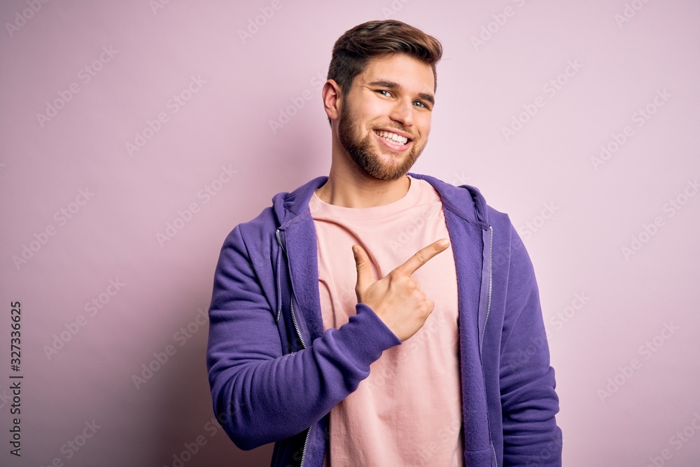 Young blond man with beard and blue eyes wearing purple sweatshirt over pink background cheerful with a smile of face pointing with hand and finger up to the side with happy and natural expression Young blond man with beard and blue eyes wearing purple sweatshirt over pink background cheerful with a smile of face pointing with hand and finger up to the side with happy and natural expression