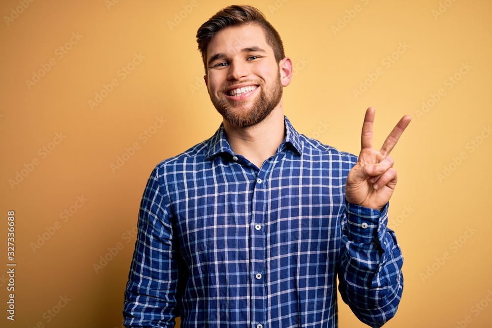 Young blond businessman with beard and blue eyes wearing shirt over yellow background showing and pointing up with fingers number two while smiling confident and happy. Young blond businessman with beard and blue eyes wearing shirt over yellow background showing and pointing up with fingers number two while smiling confident and happy.