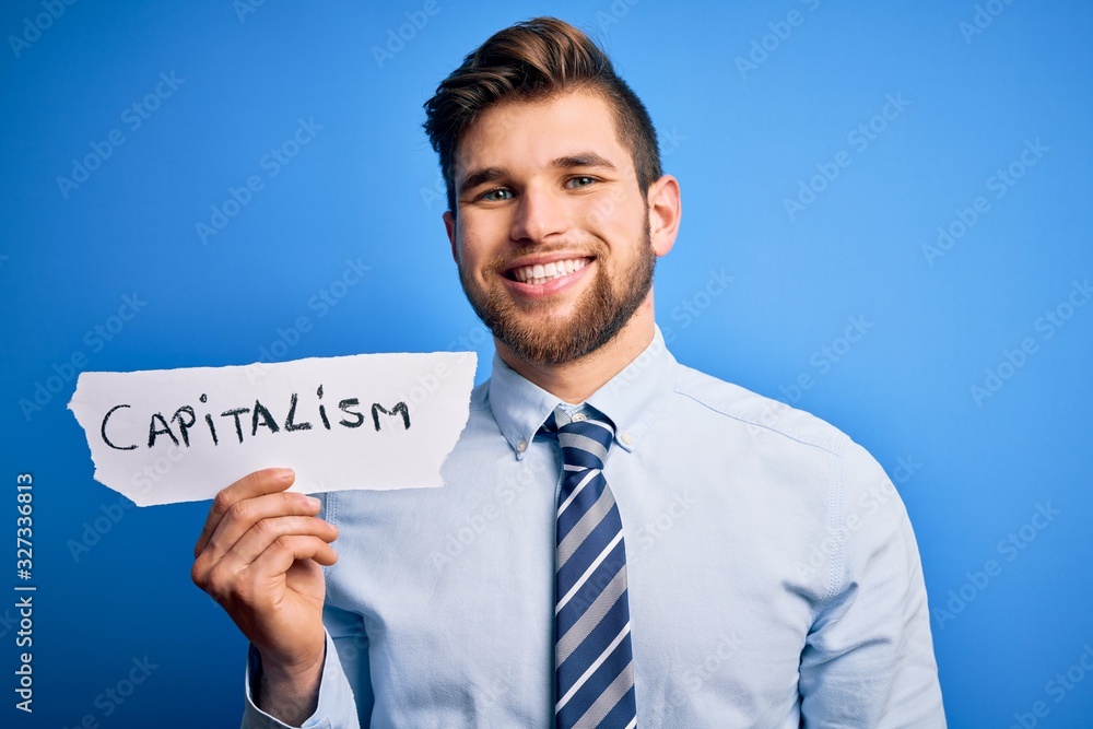 Young blond businessman with beard and blue eyes holding paper with capitalism message with a happy face standing and smiling with a confident smile showing teeth Young blond businessman with beard and blue eyes holding paper with capitalism message with a happy face standing and smiling with a confident smile showing teeth