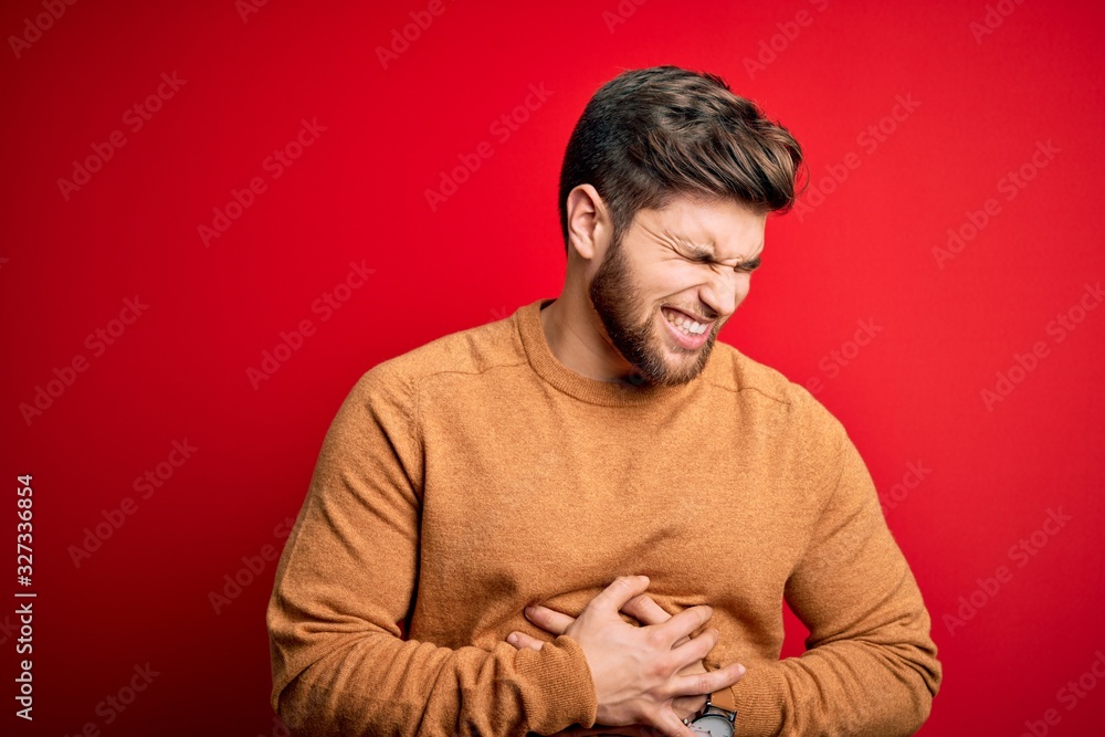 Naklejka premium Young blond man with beard and blue eyes wearing casual s over red background with hand on stomach because nausea, painful disease feeling unwell. Ache concept.