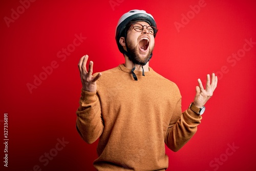 Young blond cyclist man with beard and blue eyes wearing bike helmet and glasses crazy and mad shouting and yelling with aggressive expression and arms raised. Frustration concept.