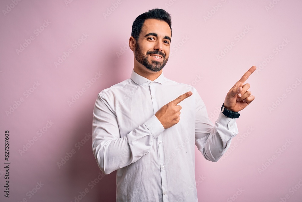Young handsome man with beard wearing casual shirt standing over pink background smiling and looking at the camera pointing with two hands and fingers to the side. Young handsome man with beard wearing casual shirt standing over pink background smiling and looking at the camera pointing with two hands and fingers to the side.