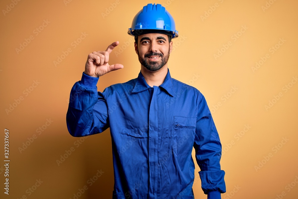 Mechanic man with beard wearing blue uniform and safety helmet over yellow background smiling and confident gesturing with hand doing small size sign with fingers looking and the camera. Measure