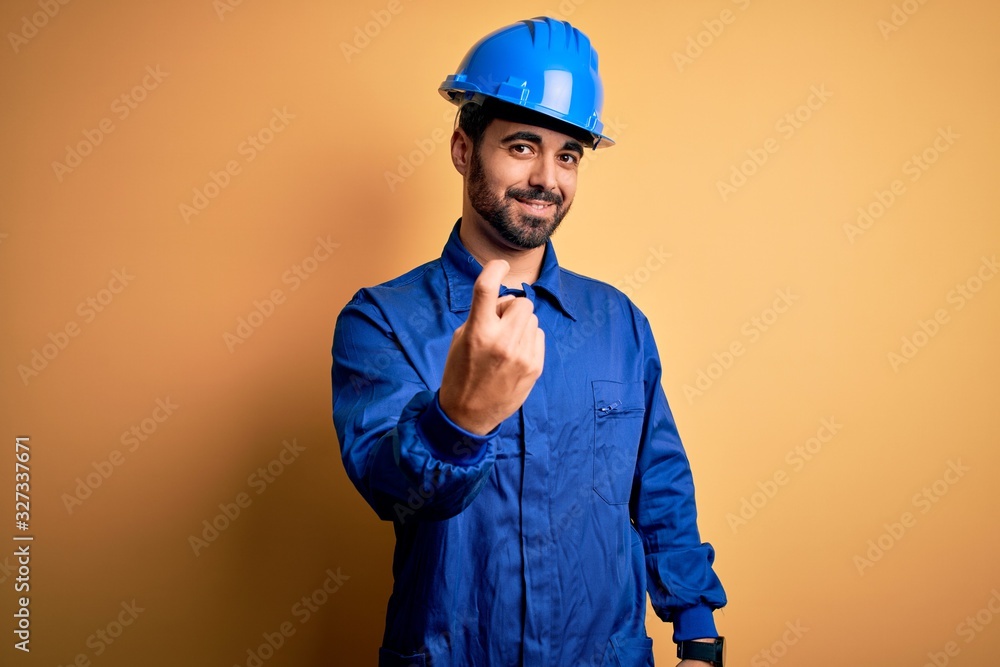 Mechanic man with beard wearing blue uniform and safety helmet over yellow background Beckoning come here gesture with hand inviting welcoming happy and smiling
