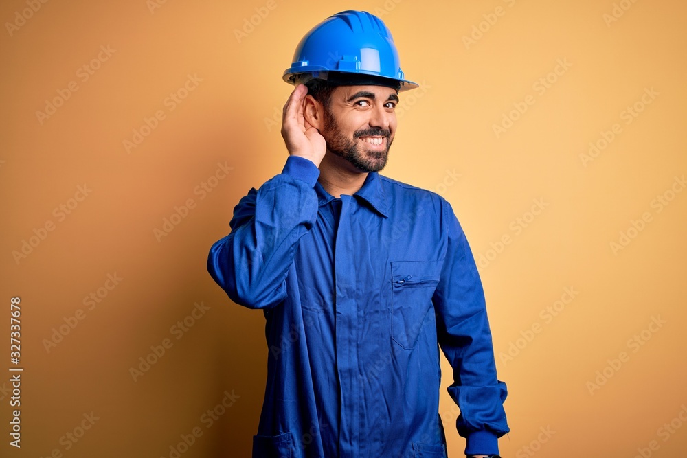 Mechanic man with beard wearing blue uniform and safety helmet over yellow background smiling with hand over ear listening an hearing to rumor or gossip. Deafness concept. Mechanic man with beard wearing blue uniform and safety helmet over yellow background smiling with hand over ear listening an hearing to rumor or gossip. Deafness concept.