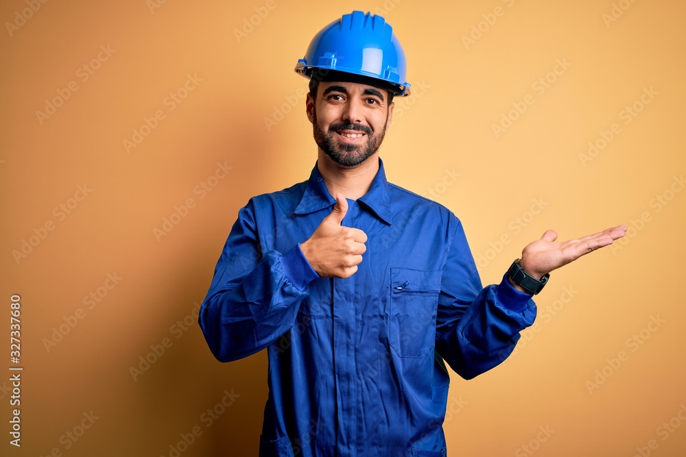 Mechanic man with beard wearing blue uniform and safety helmet over yellow background Showing palm hand and doing ok gesture with thumbs up, smiling happy and cheerful