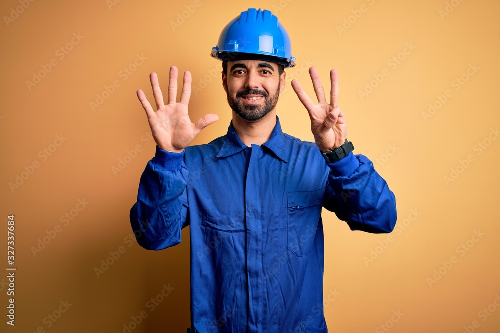 Mechanic man with beard wearing blue uniform and safety helmet over yellow background showing and pointing up with fingers number eight while smiling confident and happy.