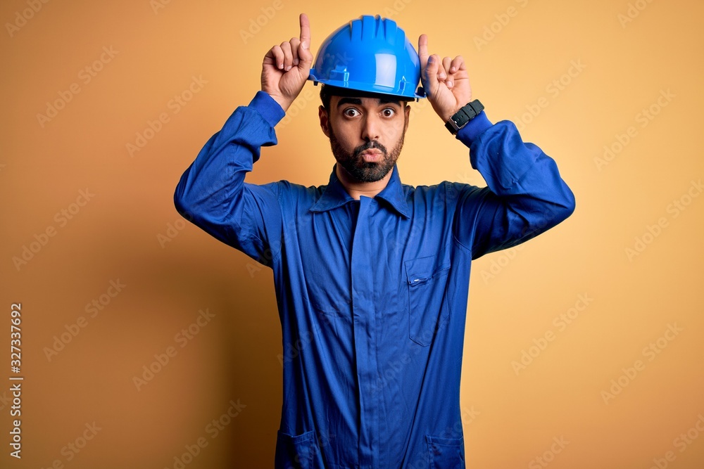 Mechanic man with beard wearing blue uniform and safety helmet over yellow background doing funny gesture with finger over head as bull horns